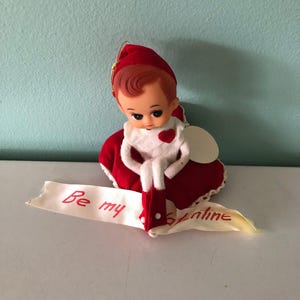 May include: A small elf doll with red hair, wearing a red hat and a white collar with a red heart accent. The doll is seated on a red fabric base and holds a white ribbon banner with the red handwritten text Be my Valentine across the front.