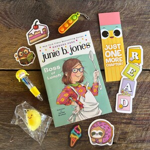 May include: A Junie B. Jones book titled Boss of Lunch, a yellow pencil-shaped bookmark with the text Just One More Chapter, a stack of blocks spelling READ, a cake sticker, a pea pod fidget, a multi-colored pen, a sloth donut sticker, and a lemon charm.