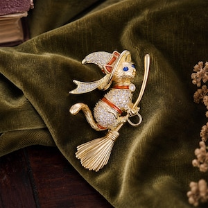 Peut inclure: Une broche chat dorée représentant un chat portant un chapeau pointu et chevauchant un balai. Le chat est orné de cristaux transparents, d'accents en émail orange et d'yeux en cristal bleu. Le balai présente une finition dorée texturée.