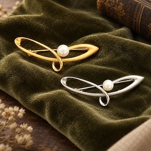 May include: Two curved metal brooches, one in a matte gold finish and one in a polished silver finish, each featuring a single white pearl accent. The brooches have an elongated, abstract shape with a looped design and a pin closure on the back.