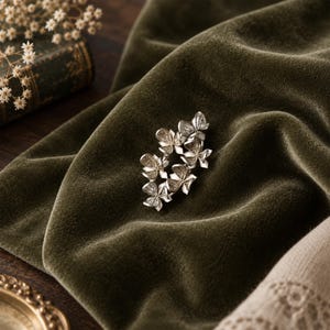 May include: A silver-toned brooch featuring a cluster of five small, detailed flower-shaped elements with textured petals. The metallic piece has a polished finish and is arranged in an elongated, organic shape, showcasing intricate floral craftsmanship.