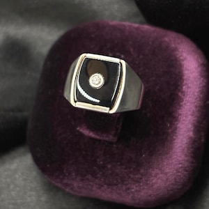 May include: A silver-toned ring featuring a rectangular black onyx inlay set into the metal band. A single, small, round, clear gemstone is embedded in the center of the black onyx stone, creating a contrast between the dark surface and the metal.