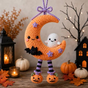 Peut inclure: Une décoration suspendue en crochet représentant un croissant de lune orange avec un visage souriant, un petit fantôme blanc, une chauve-souris noire et des étoiles violettes. La lune a deux jambes pendantes avec des bas rayés noir et violet.