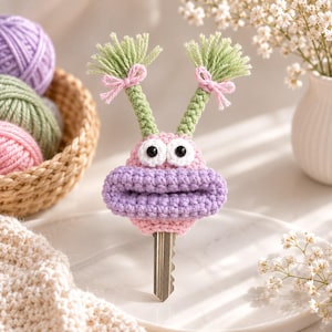 May include: A crochet key cover shaped like a pink and purple creature with two green antennae topped with light green yarn tassels and small pink bows. The creature has two large white eyes with black pupils and a wide, thick purple mouth.