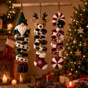 Peut inclure: Trois décorations en crochet suspendues à une tige en bois. À gauche, un gnome avec un chapeau vert et une base en forme de cadeau. Au centre, une tête de renne au-dessus d'une chaussette rouge. À droite, un sucre d'orge au-dessus d'une boule.