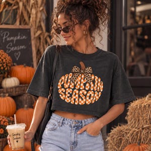 May include: A charcoal gray cropped t-shirt featuring a large orange and white checkered pumpkin graphic with a leopard print bow and the text "PUMPKIN SEASON" in a bold font. The shirt is paired with light blue denim jeans and a black shoulder bag.