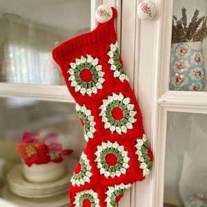 May include: A handmade crochet Christmas stocking featuring a vibrant red base colour. The design is composed of multiple square motifs, each showcasing a circular pattern in shades of dark green and light cream, arranged in a repeating grid layout.