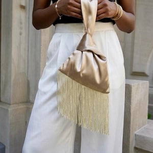 Peut inclure: Un sac à main en satin couleur champagne avec une silhouette triangulaire et une longue frange dorée fluide le long du bord inférieur. Le sac est conçu avec une anse nouée unique, créant une apparence douce et drapée.
