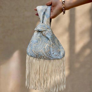 Könnte beinhalten: Eine hellblaue Handgelenktasche mit goldenem Paisley-Muster und einem langen, cremefarbenen Fransenbesatz an der Unterkante. Die Tasche hat einen geknoteten Griff und besteht aus einem glatten, satinartigen Stoff mit einem dezenten Glanz.