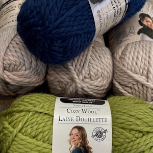 May include: A collection of five skeins of bulky yarn in navy blue, light beige, and lime green. Each skein features a paper label with the text Cozy Wool, Laine Douillette, and a photograph of a person wearing a knitted garment.