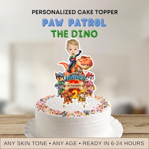 Può includere: Un topper per torta personalizzato con personaggi di Paw Patrol e un dinosauro. Il design include il volto di un bambino, il nome James e la scritta Is Turning 2 su una grafica a forma di osso. Il topper è posizionato su una torta bianca.