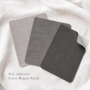 Op de afbeelding: Drie rechthoekige zelfklevende linnen reparatiepatches in lichtgrijs, middengrijs en donker antracietgrijs. Elke patch heeft een geweven textuur en afgeronde hoeken. De tekst "Self Adhesive Linen Repair Patch" staat eronder gedrukt.