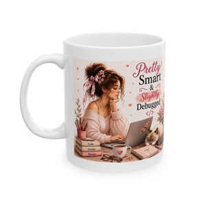 May include: A white ceramic mug featuring a graphic of a person with brown hair in a pink sweater working on a laptop. The text reads Pretty Smart & Slightly Debugged with a code symbol. A cat, books labeled Code, Create, Dream, and a mug are visible.