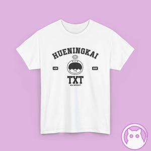 May include: A white short-sleeved t-shirt featuring a black graphic print. The design includes the text HUENINGKAI, a cartoon character head with a halo, the numbers 1408 and 2002, and the text TXT MOA UNIVERSITY in a collegiate font style.