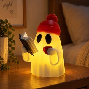May include: A glowing, off-white ghost-shaped lamp wearing a bright red knit-style beanie with a pom-pom. The ghost figure holds an open black and white book in one hand and a small red mug in the other, emitting a warm, soft yellow light.