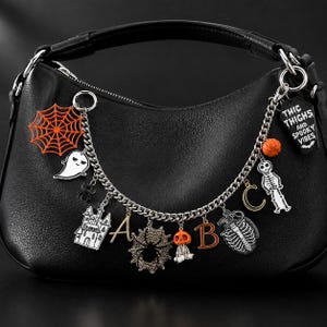 May include: A black leather handbag adorned with a silver chain featuring various charms including an orange spiderweb, a ghost, a haunted house, the letters A and B, a spider, a skull, a crescent moon, a skeleton, a pumpkin, and a coffin.