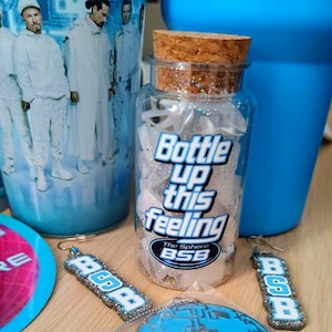 May include: A clear glass jar with a cork stopper filled with white glittery fragments. The jar features the text Bottle up this feeling and The Sphere BSB in blue and white. Two matching rectangular earrings with the letters BSB are included.