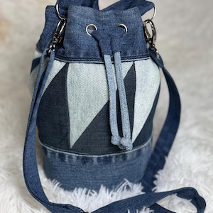 May include: A bucket bag crafted from repurposed blue denim fabric featuring a geometric star pattern in alternating light and dark wash denim panels. The bag includes a drawstring closure with silver-toned metal grommets and an adjustable strap.
