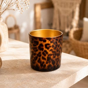 May include: A cylindrical glass candle vessel featuring a brown and black leopard print pattern on the exterior and a solid metallic gold finish on the interior. The container has a smooth, glossy surface and a wide, open top rim.