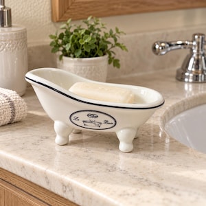 May include: A white ceramic soap dish shaped like a miniature clawfoot bathtub with a black rim. The side features an oval emblem with the text Le Bain and a small bathtub illustration. A rectangular bar of white soap rests inside the basin.
