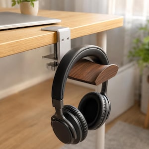 Op de afbeelding: Een zilverkleurige metalen hoofdtelefoonstandaard met een schroefklemmechanisme bevestigd aan de rand van een houten bureau. De standaard heeft een gebogen, donkerbruin houten steunstuk dat een zwarte over-ear hoofdtelefoon hangend vasthoudt.