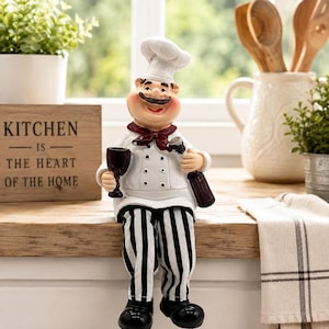 Peut inclure: Une figurine de chef décorative portant une toque blanche, une veste blanche à double boutonnage avec des boutons noirs, un foulard rouge foncé et un pantalon à rayures noires et blanches. Le chef tient un verre de vin rouge et une bouteille.