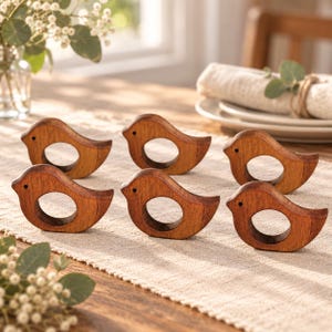 May include: A set of six bird-shaped wooden napkin rings. Each ring is crafted from brown wood with a visible grain, featuring a circular opening in the center and a small dark dot representing an eye on the head of the bird-shaped silhouette.