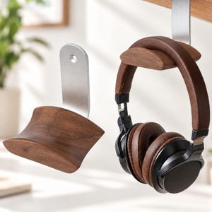 Op de afbeelding: Een hoofdtelefoonstandaard met een gebogen donkerbruine houten steun bevestigd aan een zilveren metalen beugel. Eén standaard is gemonteerd aan een oppervlak met een bruin lederen en zwarte hoofdtelefoon eraan, naast een losse standaard.