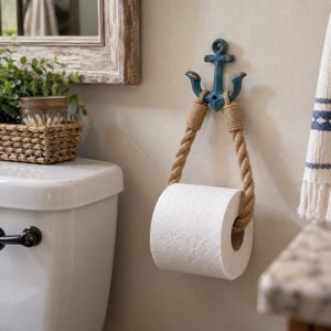 May include: A wall-mounted toilet roll holder featuring a distressed teal blue metal anchor bracket. A thick, natural-coloured jute rope is looped through the anchor, supporting a standard roll of white toilet paper hanging from the rope loop.