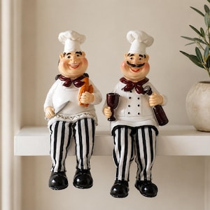 May include: Two chef figurines wearing white hats, white jackets with dark red neckerchiefs, and black and white striped trousers. One chef holds a knife and a small orange item, while the other holds a dark red wine glass and a dark brown bottle.