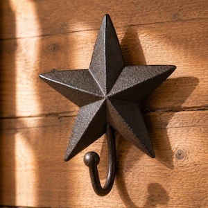 May include: A dark brown, five-pointed star wall hook made of cast metal with a textured, matte finish. The star features a central raised point with five radiating arms, and a curved hook extends from the bottom point, ending in a small ball.