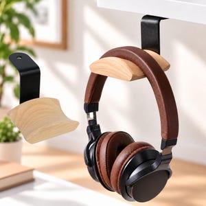 Puede incluir: Un soporte para auriculares de metal negro con una pieza de apoyo de madera clara curvada. Un soporte está montado en un estante con auriculares marrones y negros, mientras que un segundo soporte idéntico se muestra por separado a la izquierda.