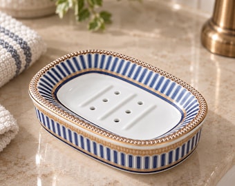 May include: An oval ceramic soap dish featuring a white center with drainage holes, surrounded by a blue and white vertical striped pattern and accented with a gold-toned decorative border along the top and bottom edges of the exterior.