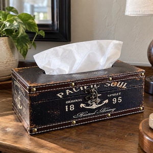May include: A rectangular tissue box cover with a distressed dark brown finish, featuring brass-tone metal studs and a front latch. The text reads PACIFIC SOUL, ORIGINAL 1895, SPIRIT, and Nautical Heritage, accompanied by a white anchor graphic.