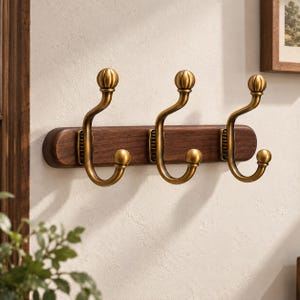 Könnte beinhalten: Eine Wandgarderobe mit einer horizontalen, dunkelbraunen Holzleiste und drei daran befestigten, messingfarbenen Metallhaken. Jeder Haken hat ein geschwungenes Design mit einem runden Abschluss oben und einem kleineren runden Ende unten.