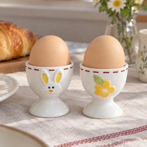 May include: Two white ceramic egg cups featuring a pedestal base and a red dashed rim. One cup displays a raised white rabbit face with yellow ears, while the other features a raised yellow flower with a green leaf. Each cup holds a brown egg.