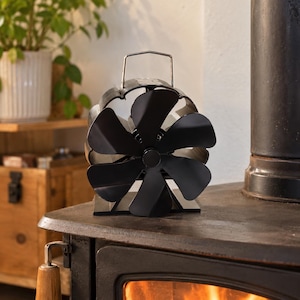 Puede incluir: Un ventilador de estufa negro alimentado por calor con seis aspas curvas y un asa de transporte de metal plateado en la parte superior. El ventilador tiene un marco circular y una base plana, colocado sobre una superficie de metal oscuro.