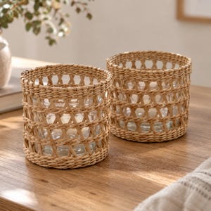 May include: Two cylindrical glass containers encased in a woven light brown natural fibre sleeve. The sleeves feature a repeating open diamond pattern with horizontal bands at the top and bottom, surrounding clear glass vessels.