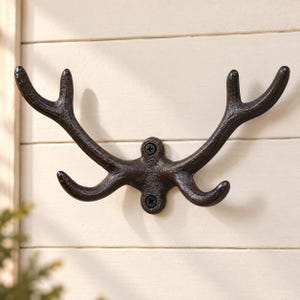 Könnte beinhalten: Ein dunkelbrauner, texturierter Wandhaken aus Metall in Form eines Hirschgeweihs. Der Haken verfügt über zwei nach oben gebogene Zinken und zwei kleinere nach unten gebogene Zinken, befestigt mit zwei sichtbaren schwarzen Schrauben.