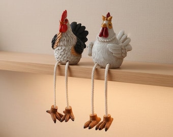 May include: Two decorative chicken figurines with textured white bodies, red combs, and long dangling rope legs ending in brown feet. The rooster features black tail feathers, while the hen wears a white beaded necklace around its neck.