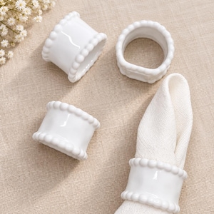 May include: Four white ceramic napkin rings with a beaded edge detail. Three of the napkin rings are empty, while one napkin ring holds a folded white fabric napkin. The rings have a smooth, glossy finish and a cylindrical shape with raised beads.