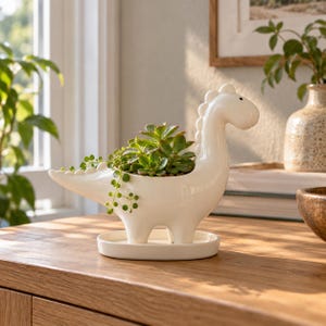 May include: A glossy white ceramic dinosaur-shaped planter featuring a long neck, a tail, and a row of rounded spikes along the back. The planter holds a green succulent plant with trailing leaves and sits on a matching oval ceramic drip tray.