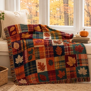 May include: A woven throw blanket featuring a patchwork design of plaid squares and various autumn leaf patterns in shades of orange, red, yellow, and dark blue. The blanket has a fringed edge and is draped over a window seat with a cream pillow.