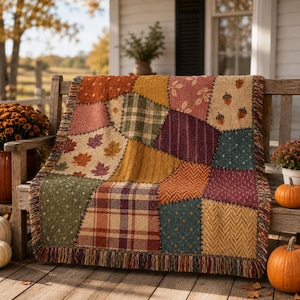 May include: A woven throw blanket featuring a patchwork design of autumn-themed patterns including maple leaves, acorns, plaid, herringbone, and polka dots in shades of orange, yellow, green, brown, and burgundy with a fringed edge on all sides.