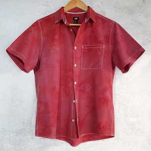 May include: A short-sleeved button-down shirt in a mottled red and pink tie-dye pattern. The shirt features a classic collar, a single chest pocket on the left side, white contrast stitching throughout, and light-coloured buttons down the front placket.
