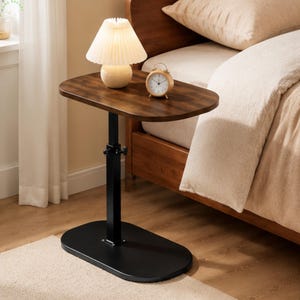 May include: A height-adjustable side table with a dark brown wood-grain oval top and a black metal base. The table features a central vertical support column with a manual adjustment knob. A small white pleated lamp and a round analog clock sit on top.
