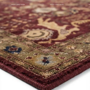 May include: A rectangular area rug featuring a deep burgundy center with intricate beige and tan floral patterns. The border is a muted olive green with blue and tan accents. The text INDIAN CARPET CENTRE is visible across the surface.