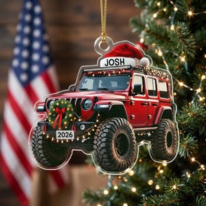 May include: A clear acrylic Christmas ornament shaped like a red off-road vehicle. The vehicle features a Santa hat on the roof with the name JOSH, a green wreath with a red bow on the front grille, and a license plate displaying the year 2026.