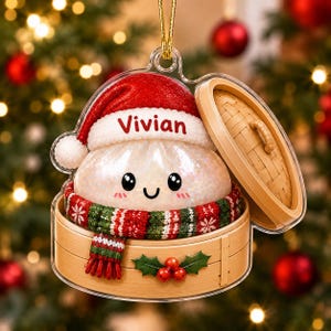 May include: A clear acrylic Christmas ornament shaped like a dumpling in a bamboo steamer. The dumpling wears a red Santa hat with the name Vivian printed on the brim and a red, white, and green knit scarf. The steamer features holly berries.