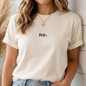Może przedstawiać: Kremowy t-shirt z krótkim rękawem z napisem no. wydrukowanym małymi czarnymi literami na klatce piersiowej. Koszulka jest zestawiona z brązowym kapeluszem typu fedora i złotym naszyjnikiem z zawieszką, noszona do jasnoniebieskich dżinsów.